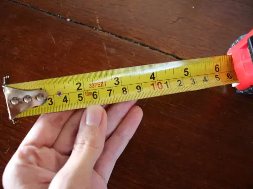 Una mano sujeta una cinta métrica amarilla sobre una mesa de madera. La cinta muestra medidas en pies y metros, demostrando para que sirve la cinta métrica: medir distancias.