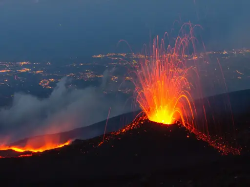 Spektakularna erupcja Etny nocą, z płynącą lawą i deszczem iskier nad oświetlonym miastem.