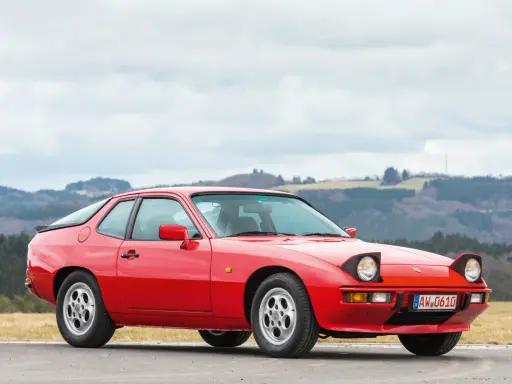 Roter Porsche 925 mit ausklappbaren Scheinwerfern steht auf einer Straße, im Hintergrund bewaldete Hügel.