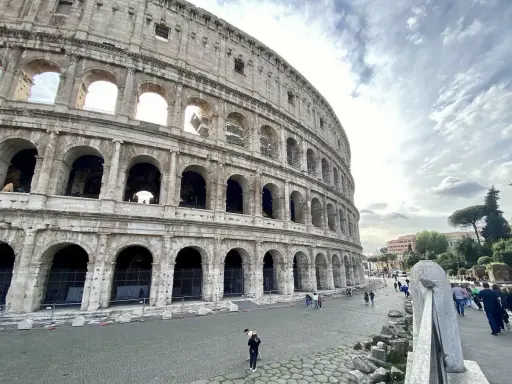 Le Colisée, monument emblématique de Rome, se dresse majestueusement sous un ciel nuageux. Des visiteurs explorent ses environs pavés.