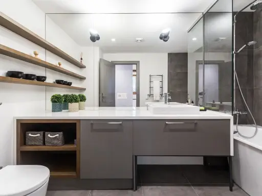 Moderno baño con ducha, bañera y lavabo sobre mueble gris. Estanterías de madera con cuencos negros. Un ejemplo de presupuesto de reforma de baño.