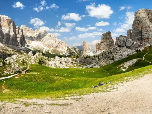 Un paesaggio mozzafiato nelle Dolomiti con le iconiche 5 torri che si ergono maestose. Un prato verde brillante invita al relax, con persone che riposano sotto il cielo azzurro.