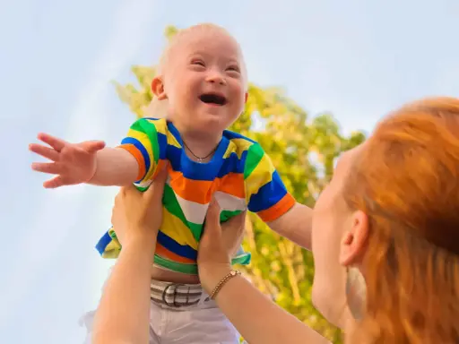 Niño con síndrome de Down, sonriente, es levantado por una persona. Sus rasgos son visibles y llenos de alegría.