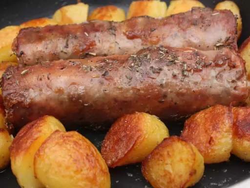 Deux andouillettes artisanales dorées, parsemées d'herbes, mijotent avec des pommes de terre rôties dans une poêle.