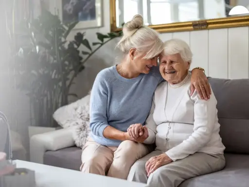 Deux femmes âgées s'enlacent tendrement sur un canapé. L'une d'elles semble visiter sa mère, une belle dame, dans sa maison en EHPAD.