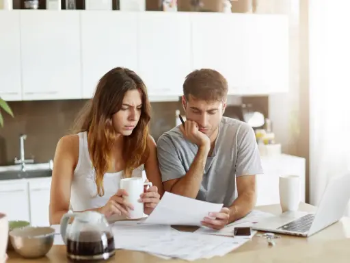 Couple pensif devant des papiers, peut-être une attestation d'assurance villégiature, dans une cuisine lumineuse.