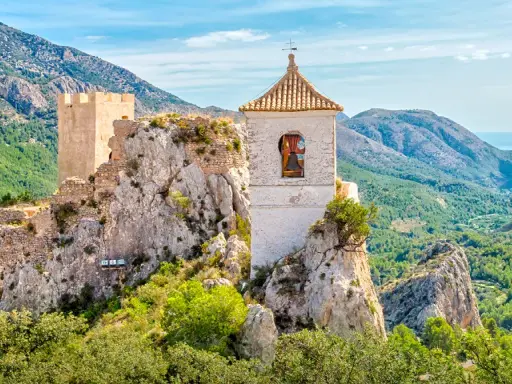 Das Castell de Guadalest thront auf einem Felsen, umgeben von grünen Bergen und dem blauen Meer in der Ferne. Ein Glockenturm ragt empor.