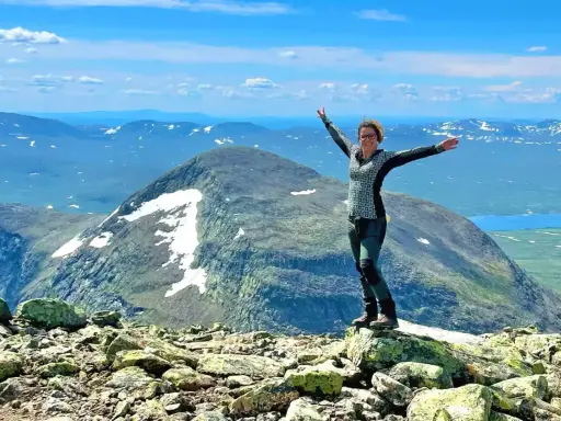 Kvinna med armarna utsträckta på en stenig topp, njuter av utsikten över fjällandskap med snöfläckar. Ett perfekt berg att bestiga!