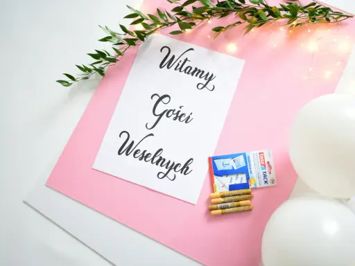 Tablica powitalna wesele z napisem "Witamy Gości Weselnych", ozdobiona gałązką zieleni i lampkami. Obok taśma, złote pisaki i białe balony.