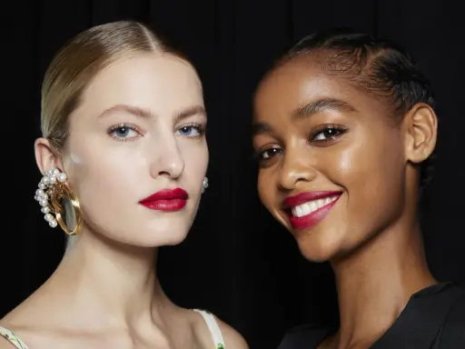 Dos modelos lucen un maquillaje para boda con labios rojos vibrantes y piel radiante. Una lleva un pendiente de perlas, la otra sonríe.