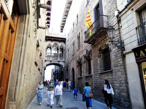 Menschen schlendern durch eine enge Gasse in der **Barcelona Altstadt**. Über ihnen spannt sich eine historische Brücke mit filigranen Bögen.