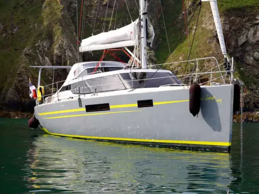 Le voilier "Bi-loup 109" gris avec des bandes jaunes, ancré près d'une falaise verdoyante.