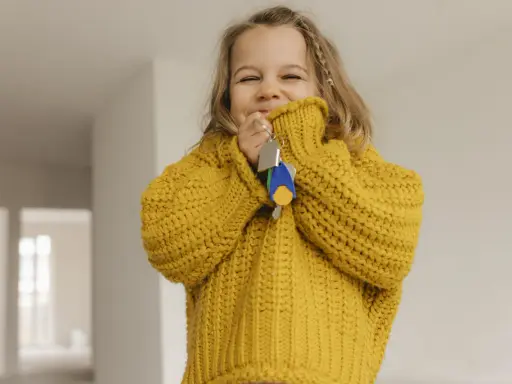 Ein fröhliches kleines Mädchen im gelben Pullover hält Schlüssel in der Hand. Die Frage "ab wann Kinder alleine lassen" beschäftigt Eltern oft, doch dieses Bild strahlt Unbeschwertheit aus.