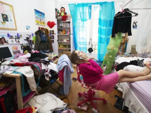 Chica en su habitación desordenada, hablando por teléfono, rodeada de ropa y objetos esparcidos.