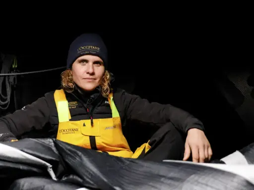 Clarisse Crémer, Seglerin im L'Occitane Sailing Team, sitzt in dunkler Kleidung und gelber Latzhose inmitten von Segeln.