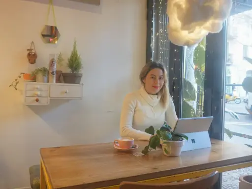 Kobieta pracuje na laptopie w kawiarni, ciesząc się kawą i roślinami. To idealne miejsce na dodatkową pracę zdalną.
