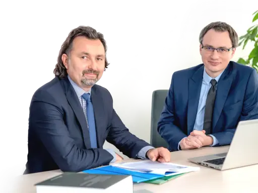 Dwóch mężczyzn w garniturach siedzi przy biurku z dokumentami i laptopem.