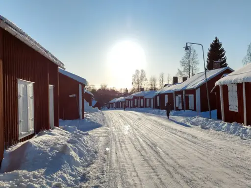 Vinterdag i kyrkbyn Gammelstad. Röda trähus med snö på taken kantar en snötäckt väg. Solen skiner starkt på en klarblå himmel.