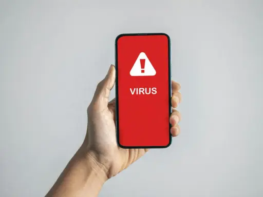 En hand håller en mobil med en röd skärm som visar en varningstriangel och texten "VIRUS". En påminnelse om hur vet man om man har virus.