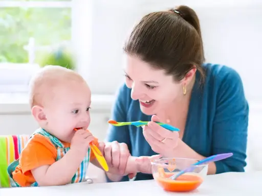Uśmiechnięta mama karmi łyżeczką 7 miesięczne dziecko, które samo próbuje jeść.