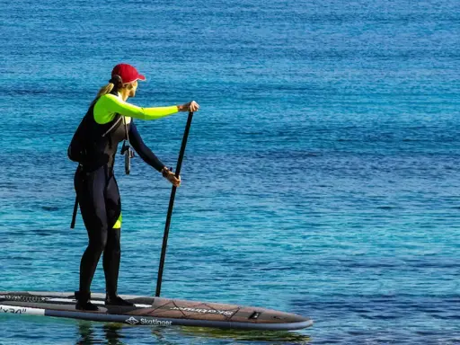 Kobieta w piance i czapce na desce sup, z wiosłem w ręku. Widać leash sup.
