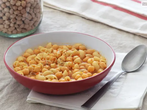 Un plato humeante de pasta e ceci, con garbanzos y macarrones en una salsa rojiza, junto a un tarro de garbanzos secos.