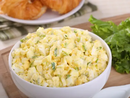 Pyszna sałatka z jajkiem, idealna na śniadanie lub lunch. Podana w białej misce, obok świeżych liści sałaty i chrupiących croissantów.