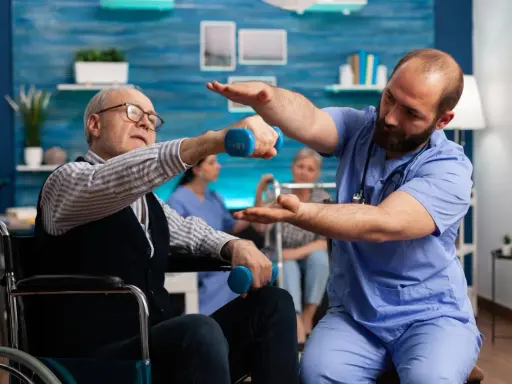 Starszy mężczyzna w wózku wykonuje ćwiczenia z hantlem pod okiem fizjoterapeuty. To kluczowy element rehabilitacji ręki po udarze.