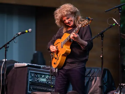 Muzyk z burzą blond włosów gra na gitarze, tworząc wspaniały śpiew w duecie z niewidocznym wokalistą.