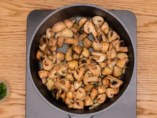 Champiñones al ajillo dorándose en una sartén, con perejil picado en un cuenco al lado.
