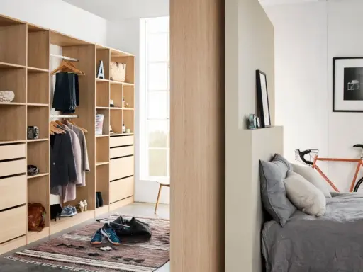 Un armario moderno y funcional, ideal para aprender como organizar un armario. Ropa colgada, cajones y estantes ordenados, con una bicicleta naranja y una cama al fondo.