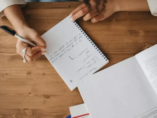 A person's hands are writing notes in a notebook. The notes mention "Theme: coronavirus", "self-isolation", and "lockdown".