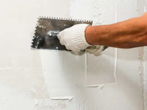 Mano enguantada aplicando estuco a una pared con una llana dentada. El proceso de estucar una pared requiere precisión.