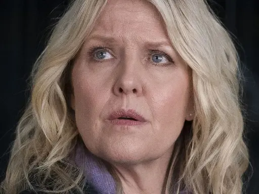 En kvinna med blont hår och blå ögon, klädd i lila, ser oroligt ut. Kanske är hon mitt i ett dramatiskt ögonblick i **shetland serie**.