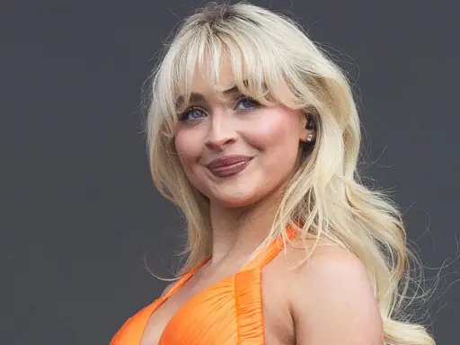 Blonde Frau mit voluminösen Blow-out-Haaren und einem strahlenden Lächeln in einem orangefarbenen Kleid.
