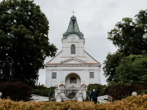 Biały kościół z zegarem na wieży, otoczony drzewami. Zastanawiasz się, jak sprawdzić do której parafii należę?