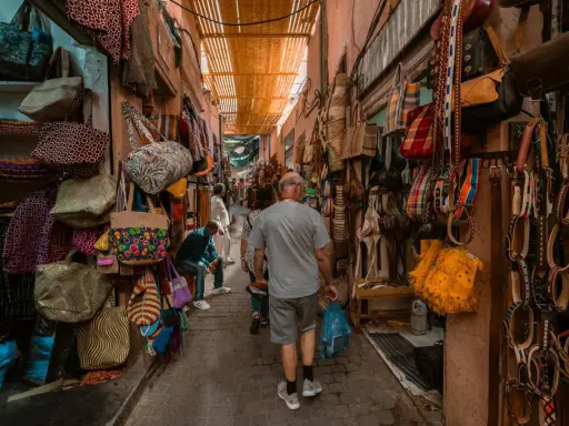 Bunte Taschen und Gürtel hängen in einem engen Gang des Marrakesch Basars. Ein Mann geht mit einer blauen Tüte durch das bunte Treiben.