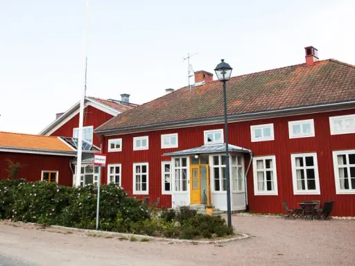 Röda trähus med vita fönster och tegeltak. En skylt visar "Stiftsgården Undersvik". Vackert beläget med natur och vatten i bakgrunden.