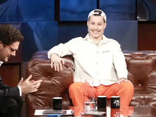 Zabson, z uśmiechem na twarzy, siedzi na kanapie w studiu. Obok niego siedzi mężczyzna w okularach.
