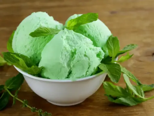 Trois boules de glace verte menthe, rappelant la saveur de la glace Get 27, garnies de feuilles de menthe fraîche dans un bol blanc sur fond de bois.