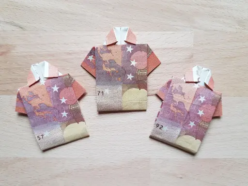Drei gefaltete Euro-Scheine in Form von Hemden liegen auf einem Holztisch. Das kunstvolle Falten von Geld zu kleinen Hemden ist eine kreative Idee.