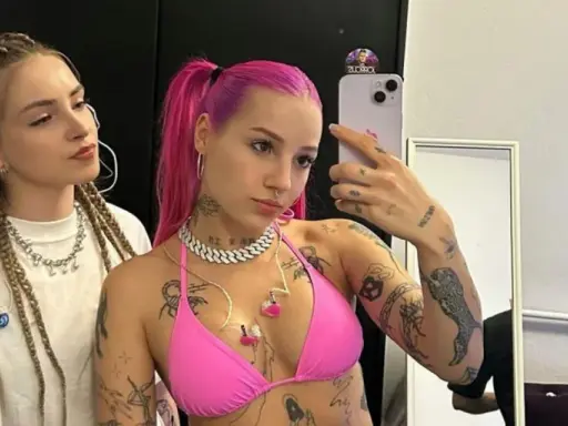Dwie młode kobiety, jedna z różowymi włosami i tatuażami, druga z warkoczami, pozują do selfie.