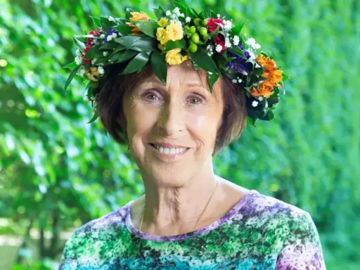 Eva Rydberg ler glatt med en blomsterkrans på huvudet, omgiven av grönska.