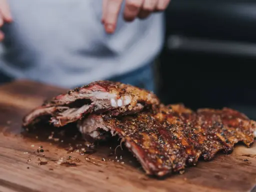 Saftige, gewürzte 321 Ribs liegen auf einem Holzbrett. Hände im Hintergrund bereiten sie für den Genuss vor.