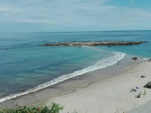 La **playa de Pechón** se extiende con arena dorada y aguas turquesas. Unas rocas emergen del mar, mientras algunas personas disfrutan del sol y el mar.