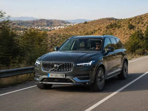 Volvo XC90 oscuro, uno de los suv mas fiables, circulando por una carretera rural con colinas verdes de fondo.