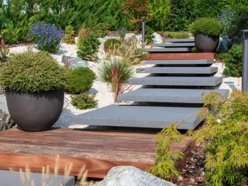 Escalier moderne en pierre et bois menant à un jardin paysager sans entretien, avec des graminées ornementales et des arbustes.