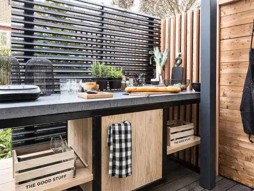 Cucina all'aperto fai da te con piano in cemento, pane, verdure e utensili. Un grembiule nero è appeso a una parete di legno.