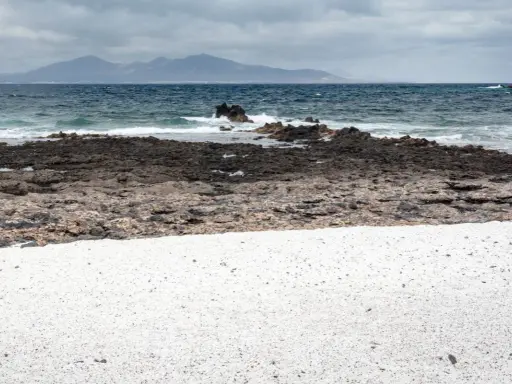 Arena blanca y rocas volcánicas en la playa Palomitas, Fuerteventura. El mar azul choca contra las rocas, con montañas al fondo.