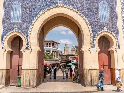 Prachtvolles Tor in Fes Stadt mit blau-goldenen Mustern. Menschen beleben den Platz dahinter.